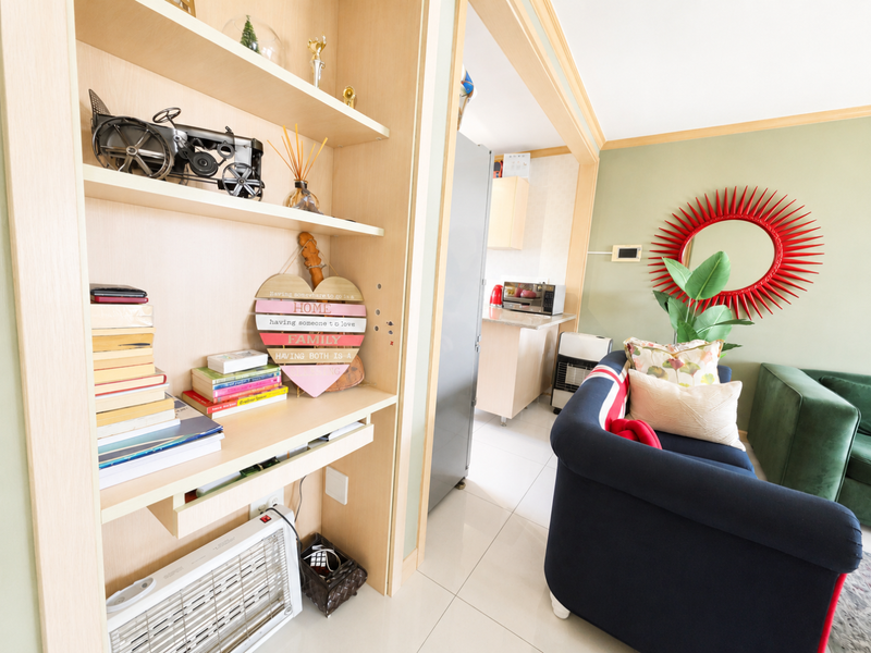 An open-plan living area featuring a built-in wooden shelving unit, tiled flooring, and a view through to the adjacent kitchen space.