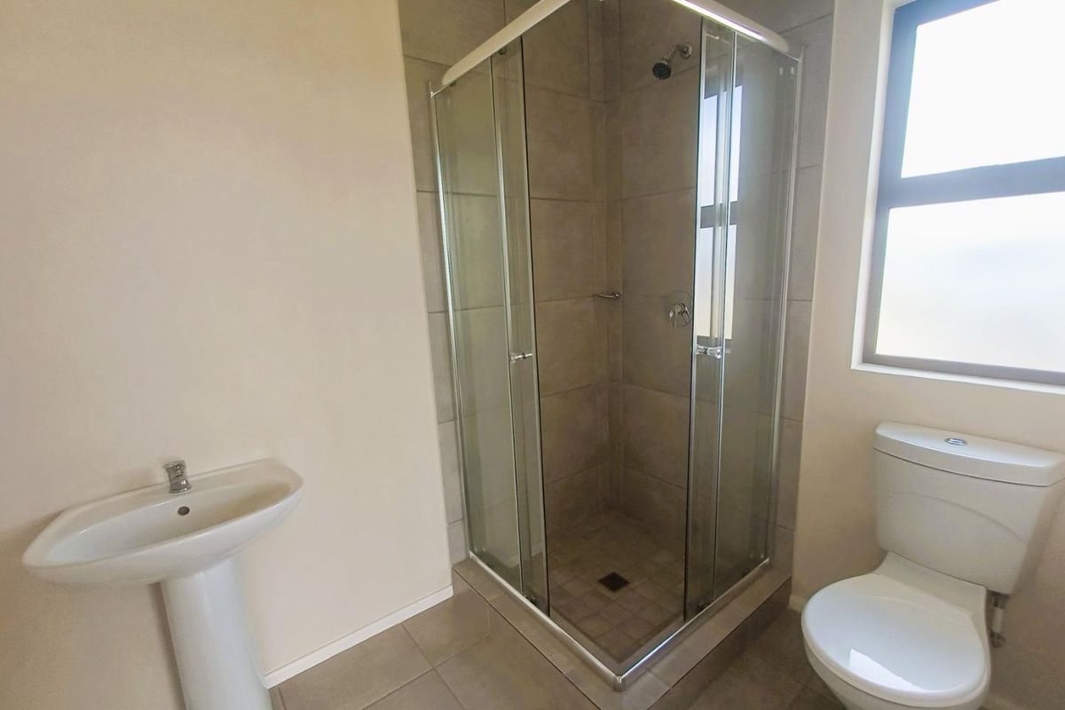 A clean, functional bathroom featuring a corner glass shower enclosure, pedestal basin, and a standard toilet with neutral floor and wall tiling.