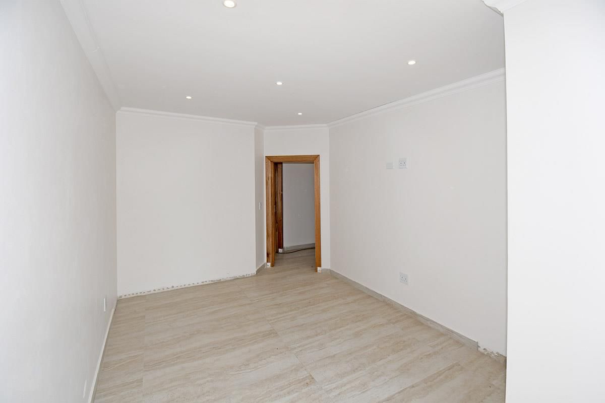 A bright, empty living area featuring neutral tiled flooring, recessed ceiling lighting, and a wooden door frame leading to an adjacent room.