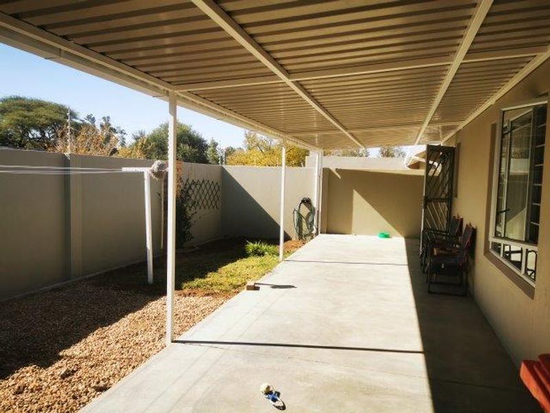 A spacious covered patio area featuring concrete flooring and a metal roof structure, overlooking a private garden with perimeter boundary walls.