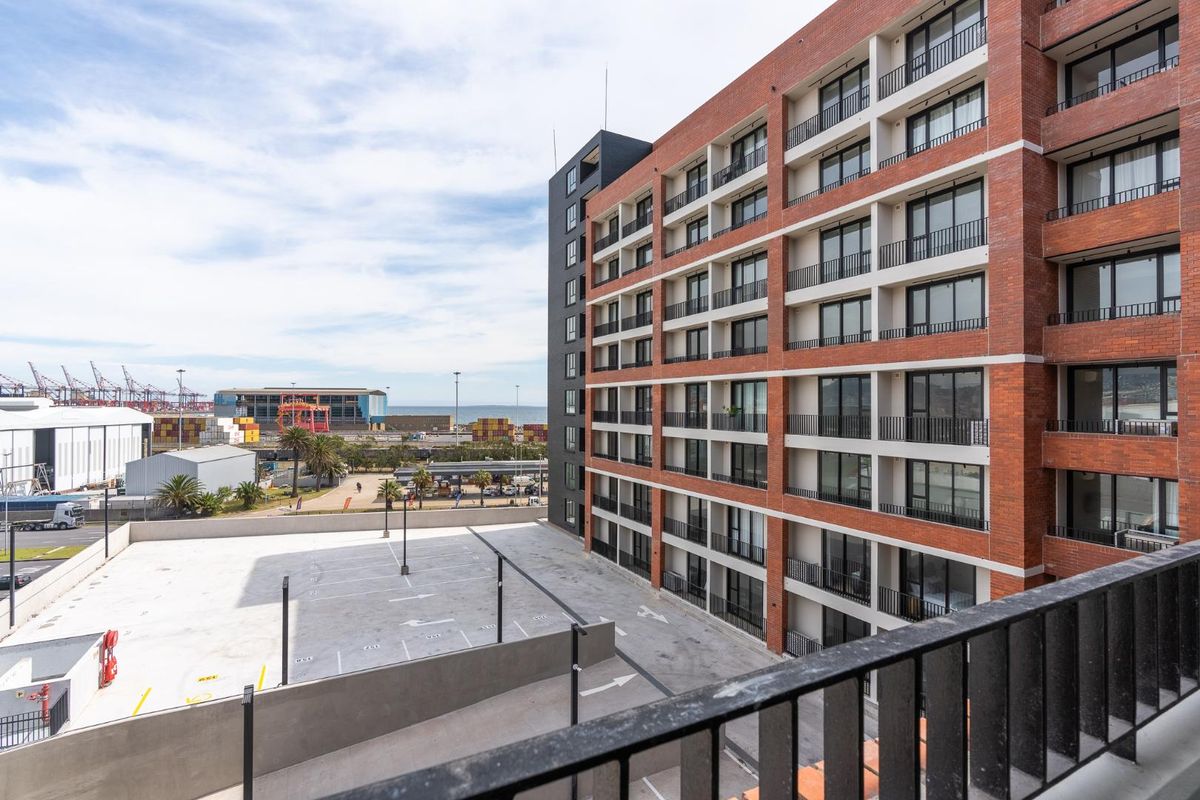 Exterior view of a modern brick apartment building featuring multiple balconies and an adjacent paved parking area with industrial surroundings.