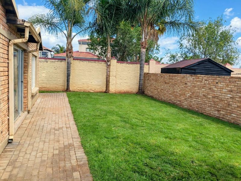 A private rear garden featuring a paved walkway, well-maintained lawn, and perimeter brick boundary walls with established palm trees.