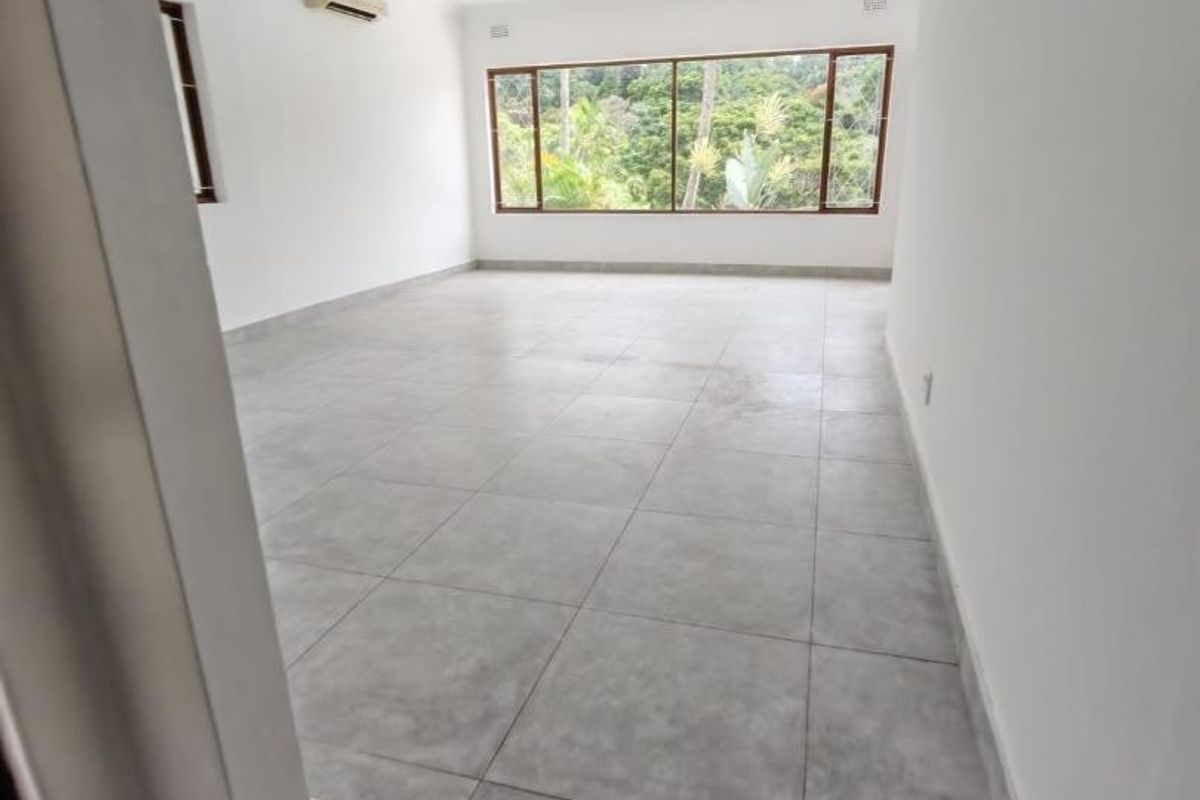 A bright, spacious, and empty living area featuring large windows with a scenic view, neutral tiled flooring, and a wall-mounted air conditioning unit.