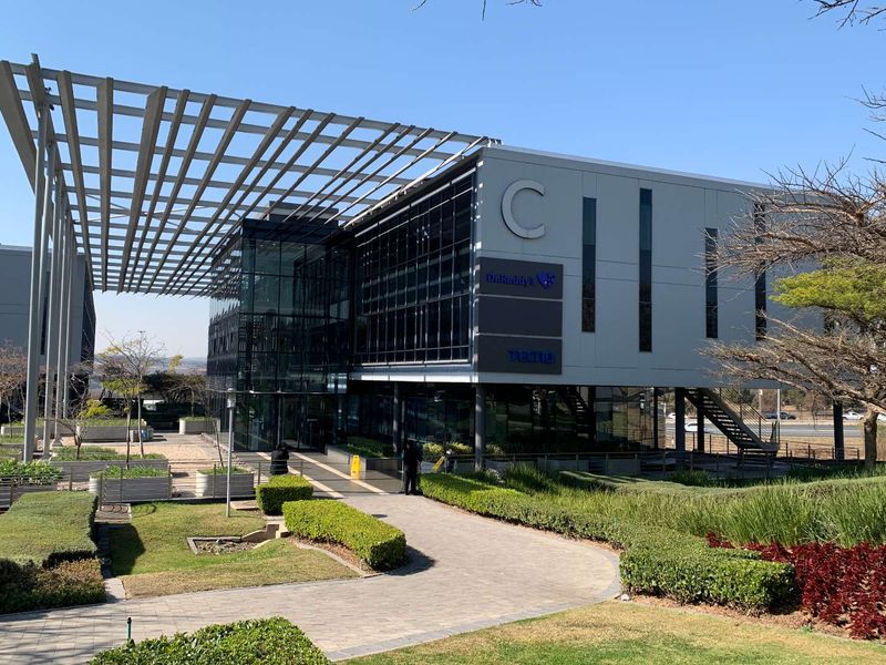 Modern commercial office building featuring a striking glass facade, architectural steel pergola, and well-maintained landscaped grounds with paved walkways.