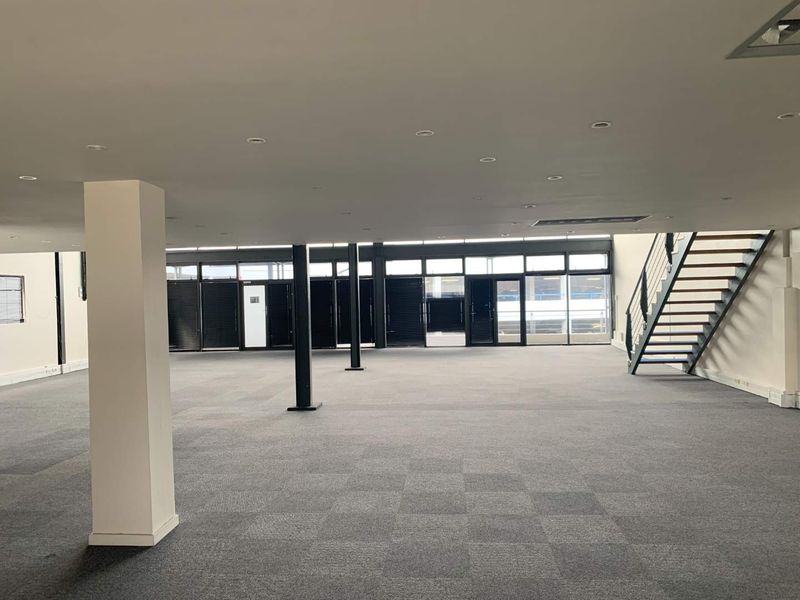 A spacious, open-plan office area featuring carpeted flooring, structural columns, recessed lighting, and a modern metal staircase leading to an upper level.