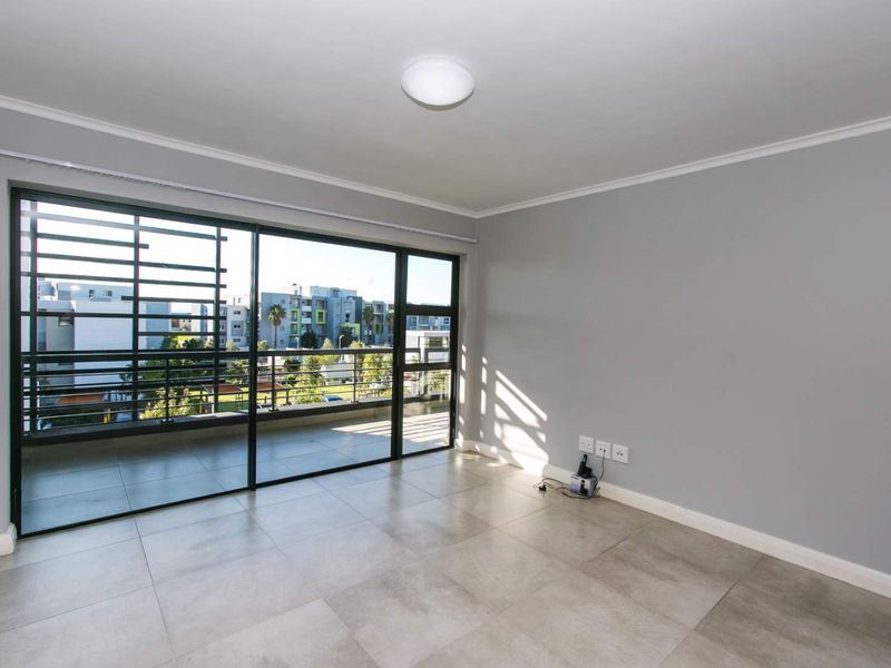A bright, empty living room featuring large sliding glass doors that open onto a balcony, with neutral tiled flooring and grey walls.