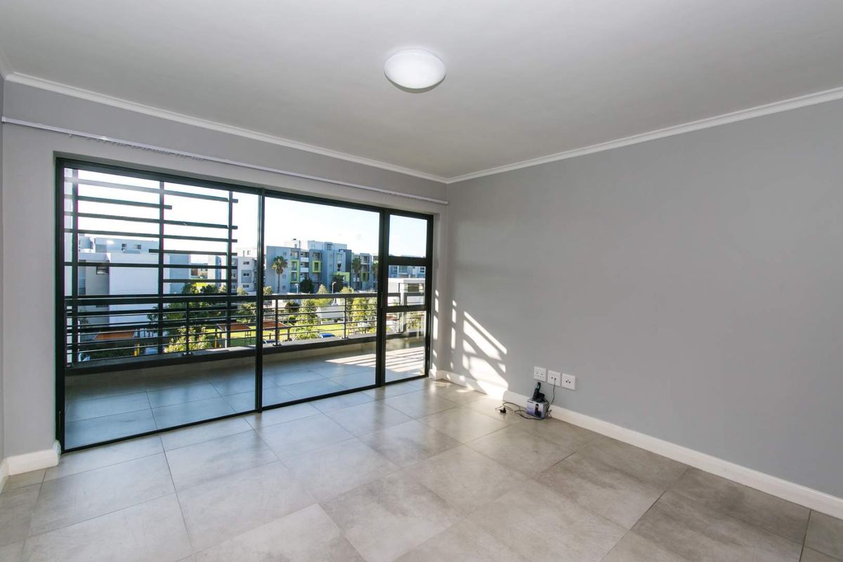 A bright, empty living room featuring large sliding glass doors that open onto a balcony, with neutral tiled flooring and grey walls.