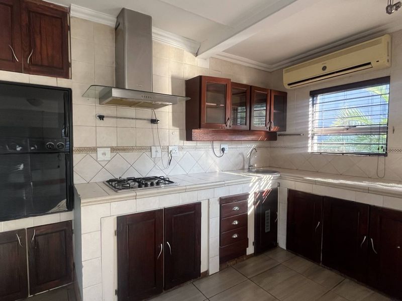 A functional kitchen featuring dark wood cabinetry, tiled countertops, a gas hob with extractor, built-in oven, and a wall-mounted air conditioning unit.