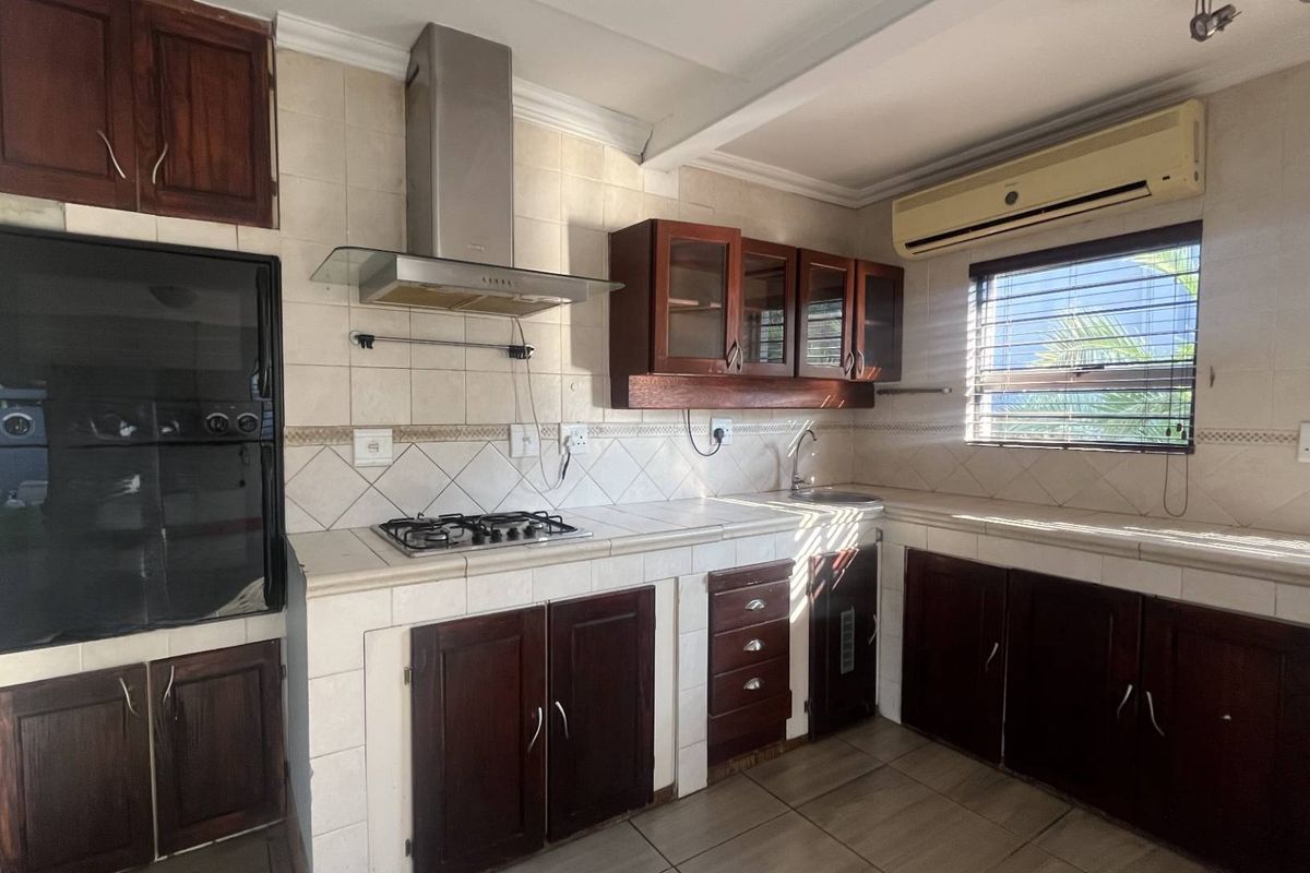 A functional kitchen featuring dark wood cabinetry, tiled countertops, a gas hob with extractor, built-in oven, and a wall-mounted air conditioning unit.