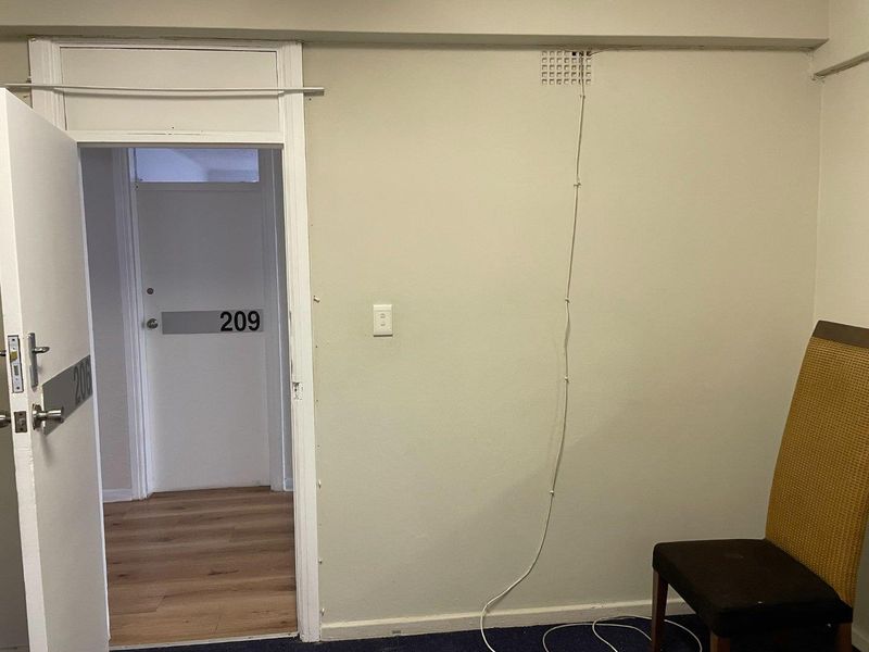 An office space featuring laminate flooring and a doorway leading to a corridor, with visible surface-mounted wiring and dated finishes.