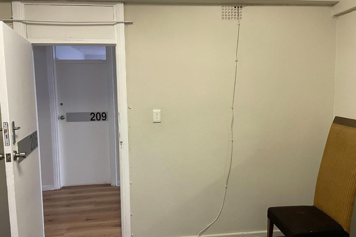 An office space featuring laminate flooring and a doorway leading to a corridor, with visible surface-mounted wiring and dated finishes.