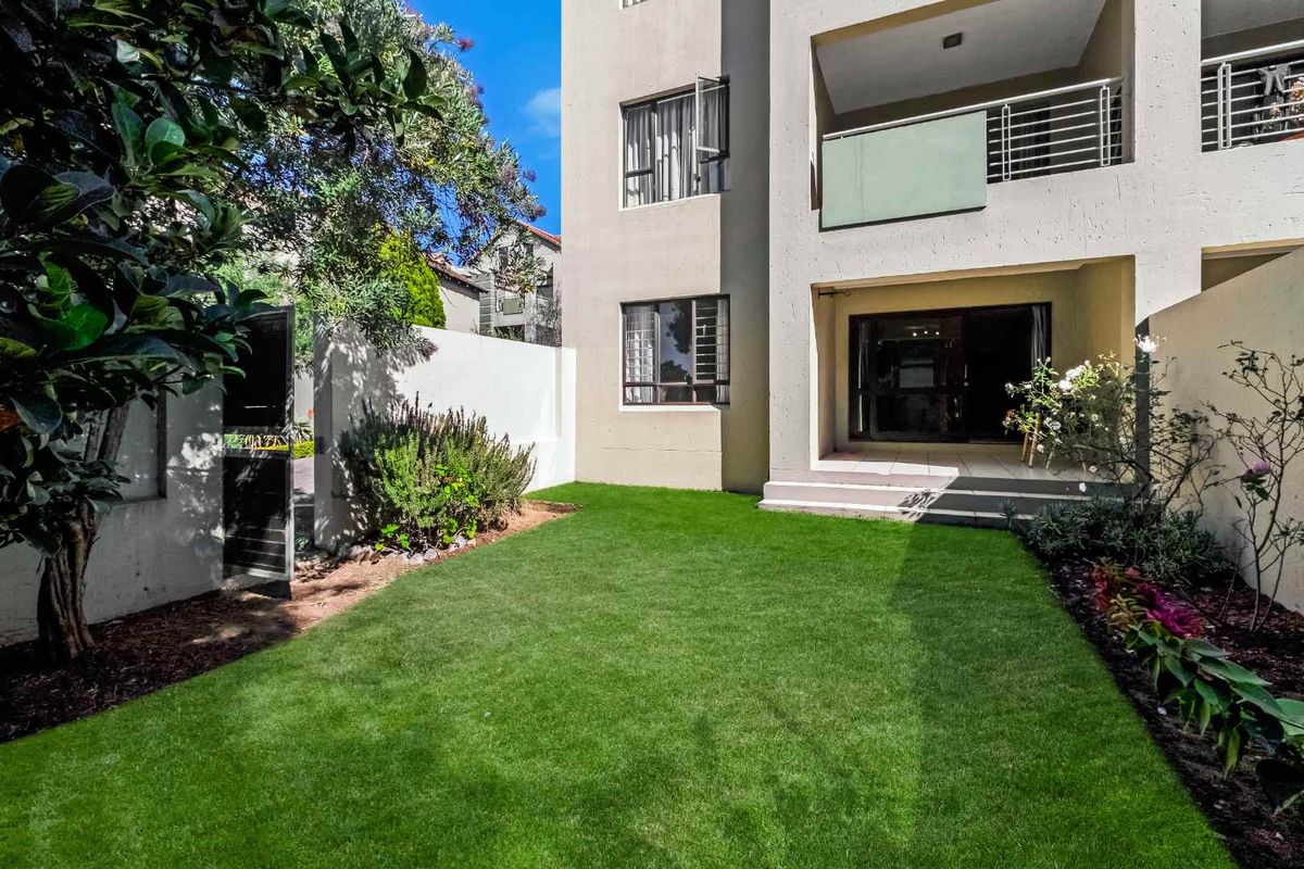 A private rear garden featuring a well-maintained lawn, landscaped borders, and a covered patio area attached to the apartment building.