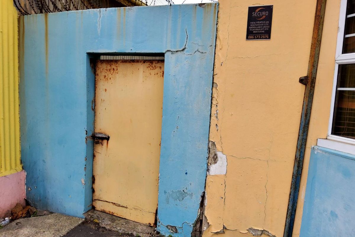 Exterior view of a service entrance featuring a weathered metal door, security signage, and perimeter wall with barbed wire.