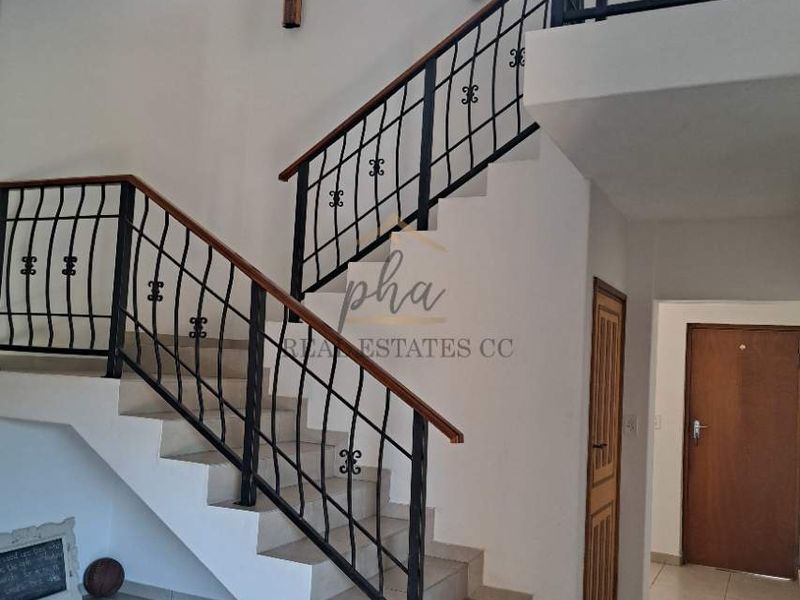 A bright, open-plan hallway featuring a staircase with a decorative wrought iron balustrade, wooden handrail, and tiled flooring.