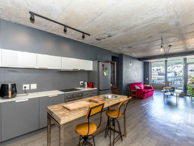 Modern open-plan living area featuring an industrial-style exposed concrete ceiling, sleek kitchen cabinetry with integrated appliances, and seamless flow to a balcony with city views.