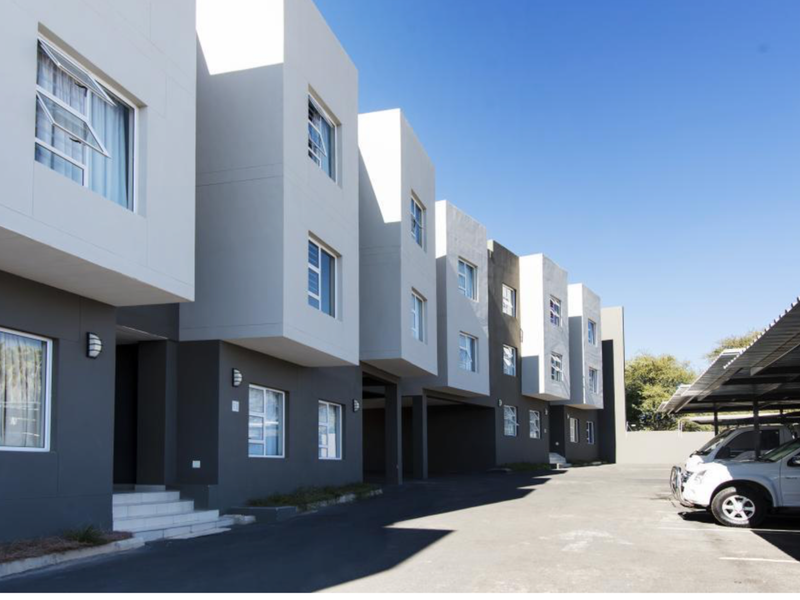 Modern townhouse complex exterior featuring contemporary architectural design, paved driveway, and covered parking bays under a clear blue sky.