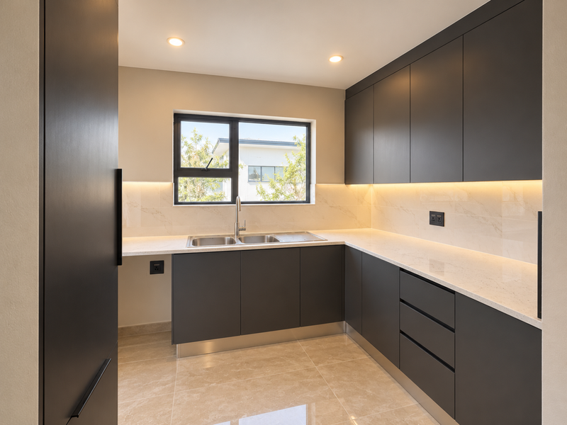 A modern, minimalist kitchen featuring sleek dark cabinetry, light stone countertops, a double sink, and integrated under-cabinet lighting in a clean, neutral space.