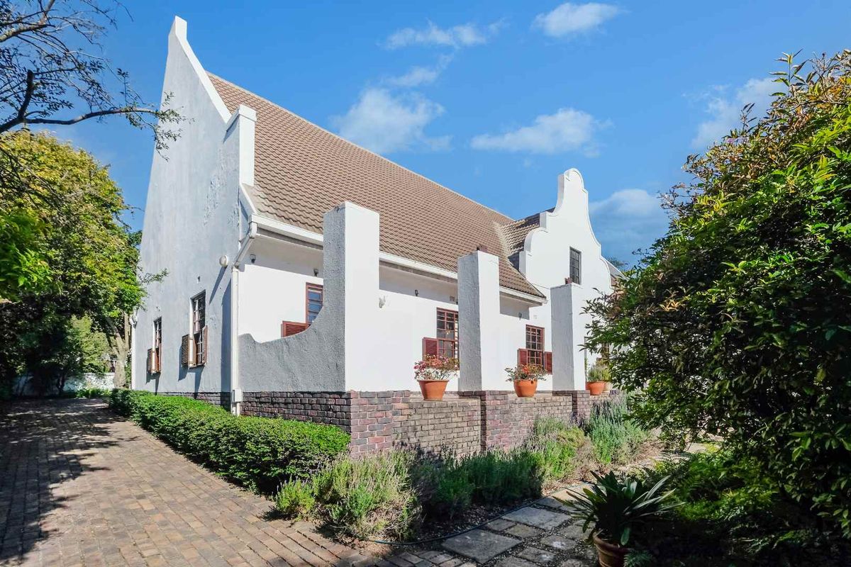 A side view of a Cape Dutch style house featuring white gabled walls, a tiled roof, and a brick-paved walkway surrounded by established garden landscaping.