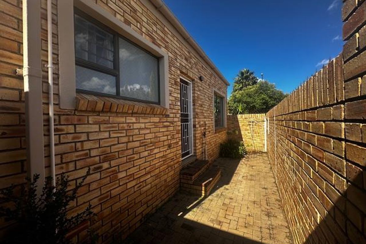 A side exterior view of the townhouse featuring face-brick walls, paved walkway, and a security gate entrance.