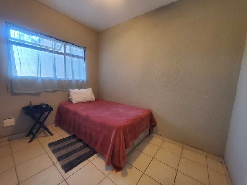 A simple bedroom featuring tiled flooring and a large window, providing natural light. The space requires cosmetic updates to the wall finishes.