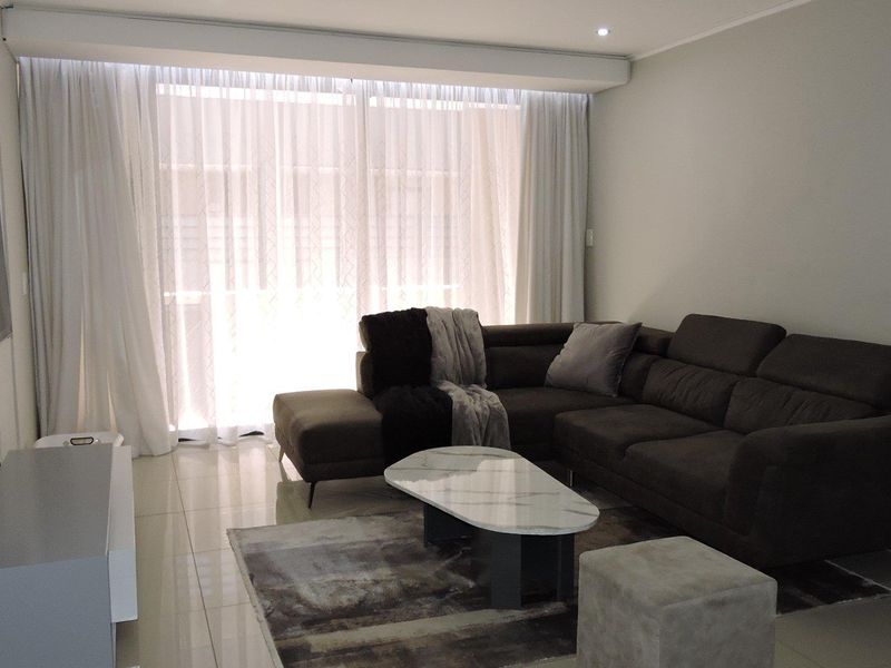 A well-lit living room featuring neutral tiled flooring, recessed ceiling lighting, and large windows dressed with floor-to-ceiling curtains.