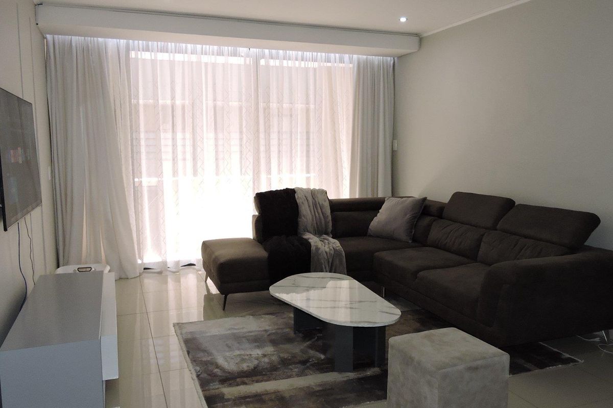 A well-lit living room featuring neutral tiled flooring, recessed ceiling lighting, and large windows dressed with floor-to-ceiling curtains.