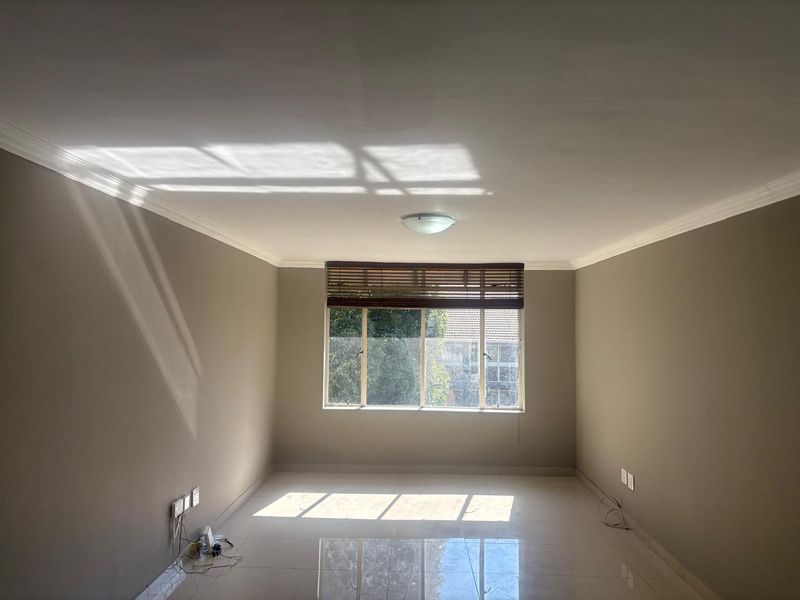 A bright, empty living room featuring neutral-toned walls, tiled flooring, and a large window with fitted blinds, offering a clean and versatile living space.