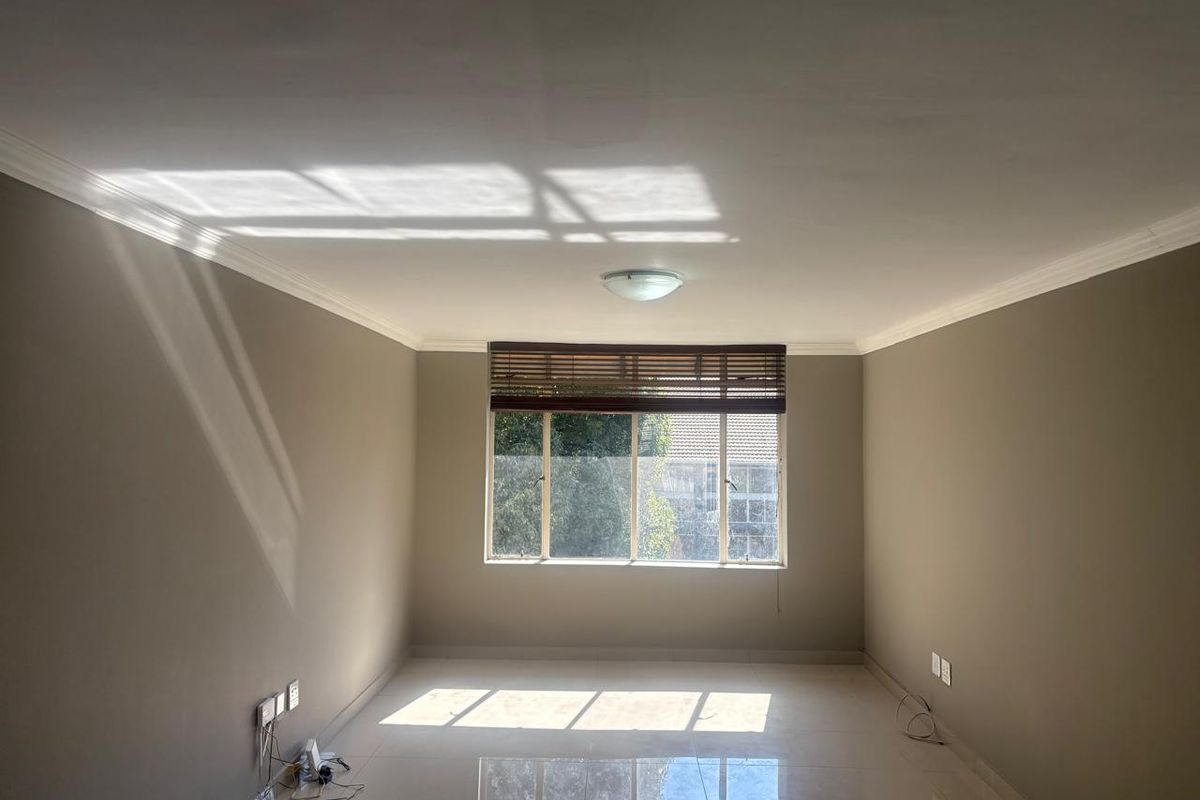 A bright, empty living room featuring neutral-toned walls, tiled flooring, and a large window with fitted blinds, offering a clean and versatile living space.