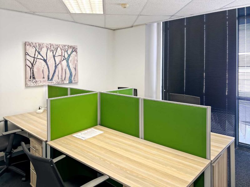 A functional office space featuring built-in workstation partitions, suspended ceiling tiles, and large windows with venetian blinds for natural light control.