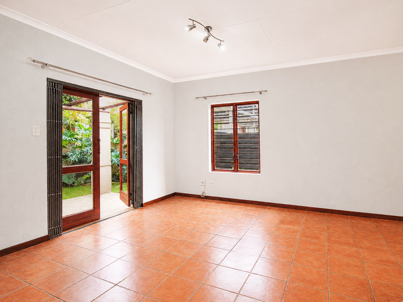 A bright, empty living room featuring terracotta floor tiles, wooden folding doors leading to a patio, and security bars on the window.