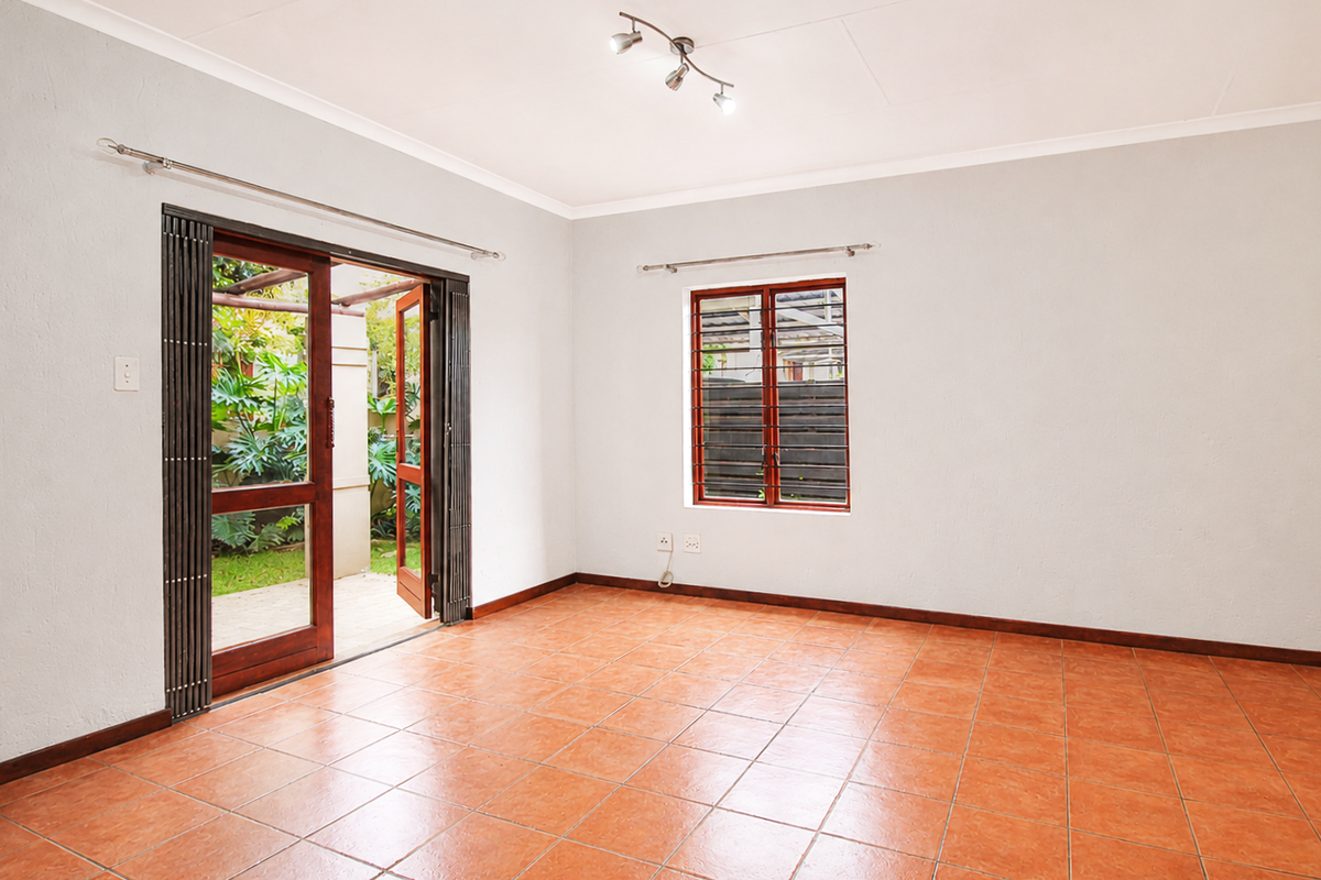A bright, empty living room featuring terracotta floor tiles, wooden folding doors leading to a patio, and security bars on the window.