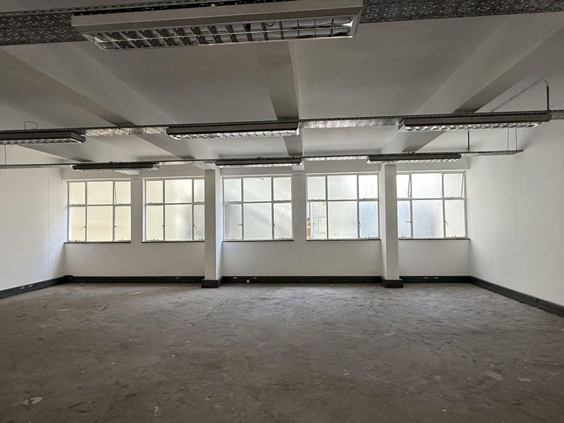 A spacious, open-plan office area featuring exposed ceiling infrastructure, industrial-style lighting, and large windows providing ample natural light.