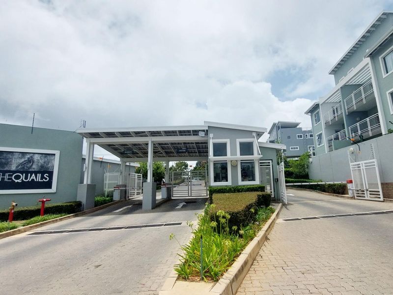 The secure entrance to The Quails apartment complex, featuring a manned gatehouse, automated boom gates, and perimeter electric fencing.