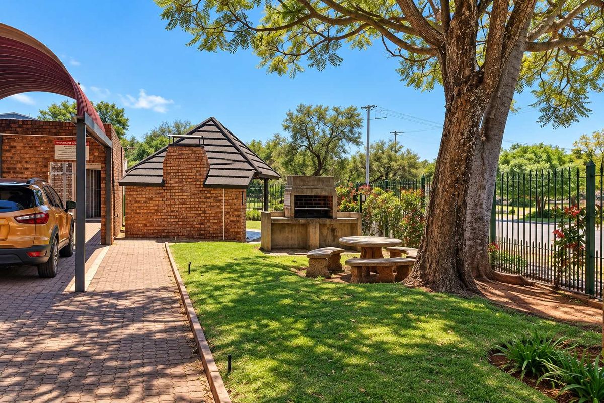 Communal outdoor area featuring a brick braai, paved driveway, and established trees within a secure fenced complex.