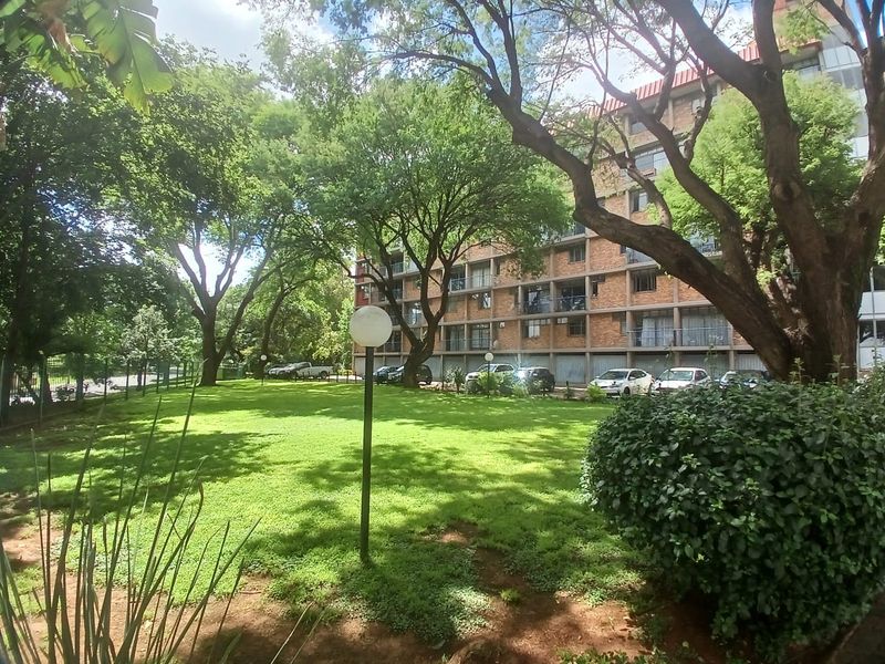 A view of the apartment building exterior featuring a well-maintained communal garden area with mature trees and brick facade architecture.