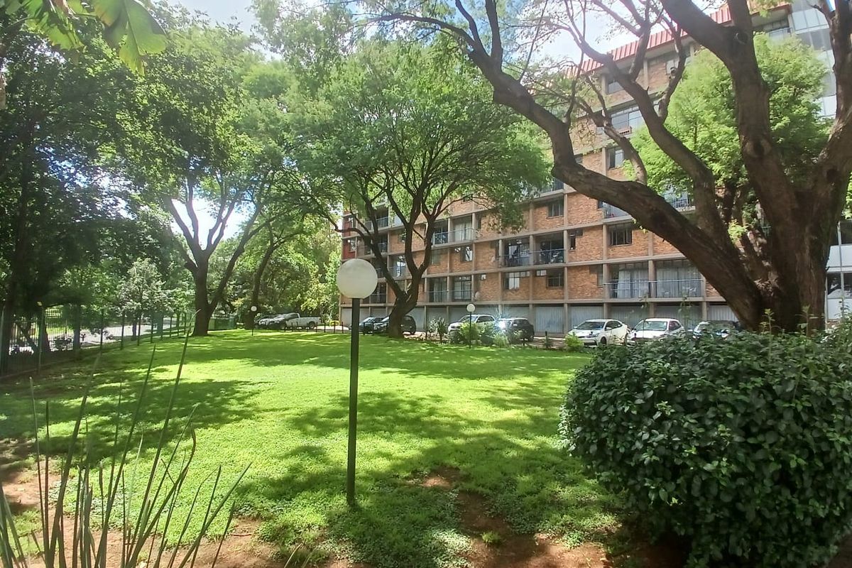 A view of the apartment building exterior featuring a well-maintained communal garden area with mature trees and brick facade architecture.