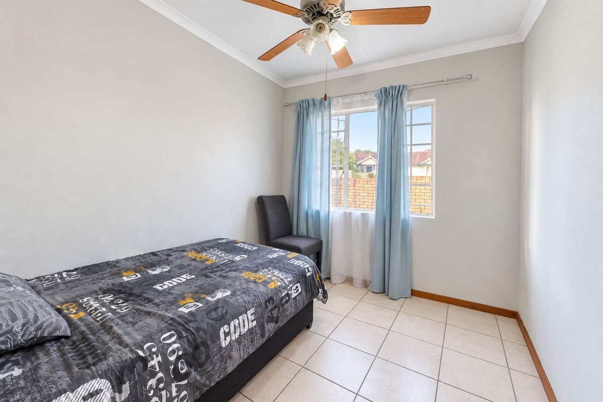A functional bedroom featuring tiled flooring, a ceiling fan, and a window with security bars, providing a clean and simple living space.