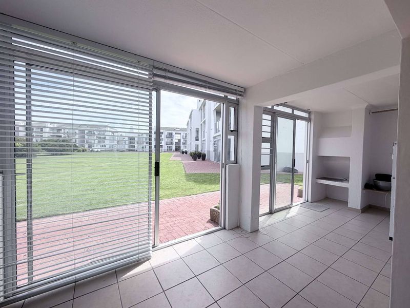 A spacious covered patio featuring tiled flooring, a built-in braai, and large sliding glass doors that open onto a communal garden area.