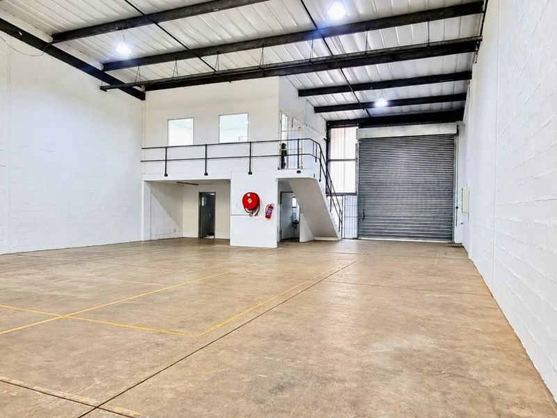 Spacious industrial warehouse unit featuring high ceilings with exposed steel trusses, a roller shutter door, concrete flooring, and a functional mezzanine level.