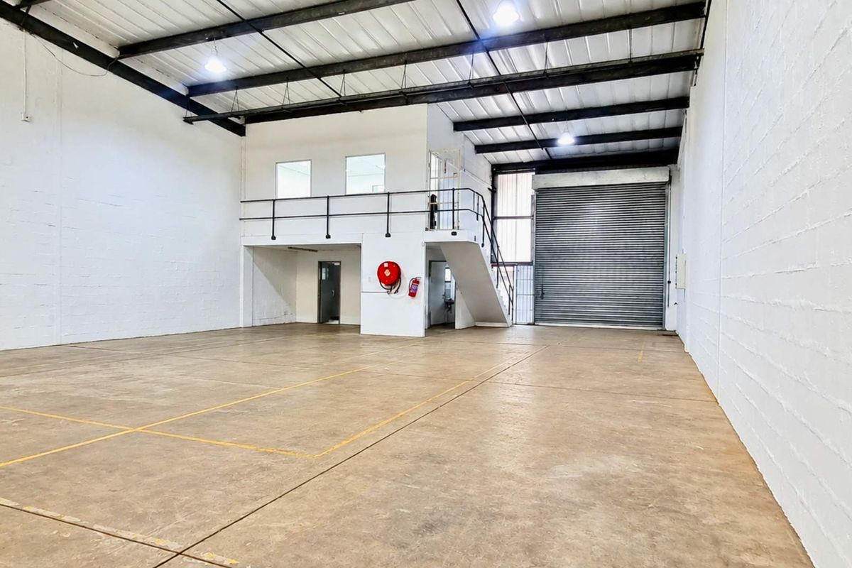 Spacious industrial warehouse unit featuring high ceilings with exposed steel trusses, a roller shutter door, concrete flooring, and a functional mezzanine level.