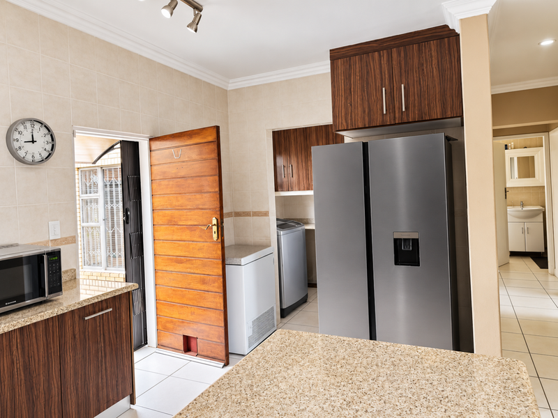 A functional scullery area featuring wood-finish cabinetry, granite countertops, and tiled surfaces, with direct access to an outdoor space and a view into a bathroom.