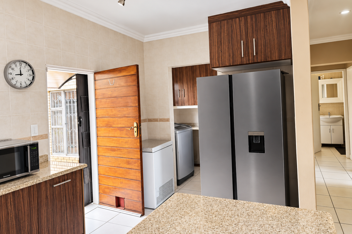 A functional scullery area featuring wood-finish cabinetry, granite countertops, and tiled surfaces, with direct access to an outdoor space and a view into a bathroom.