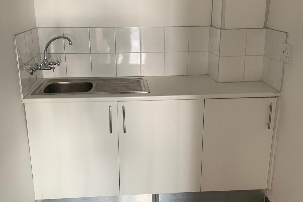 A compact kitchenette area featuring white built-in cabinetry, a stainless steel sink, and a tiled backsplash.