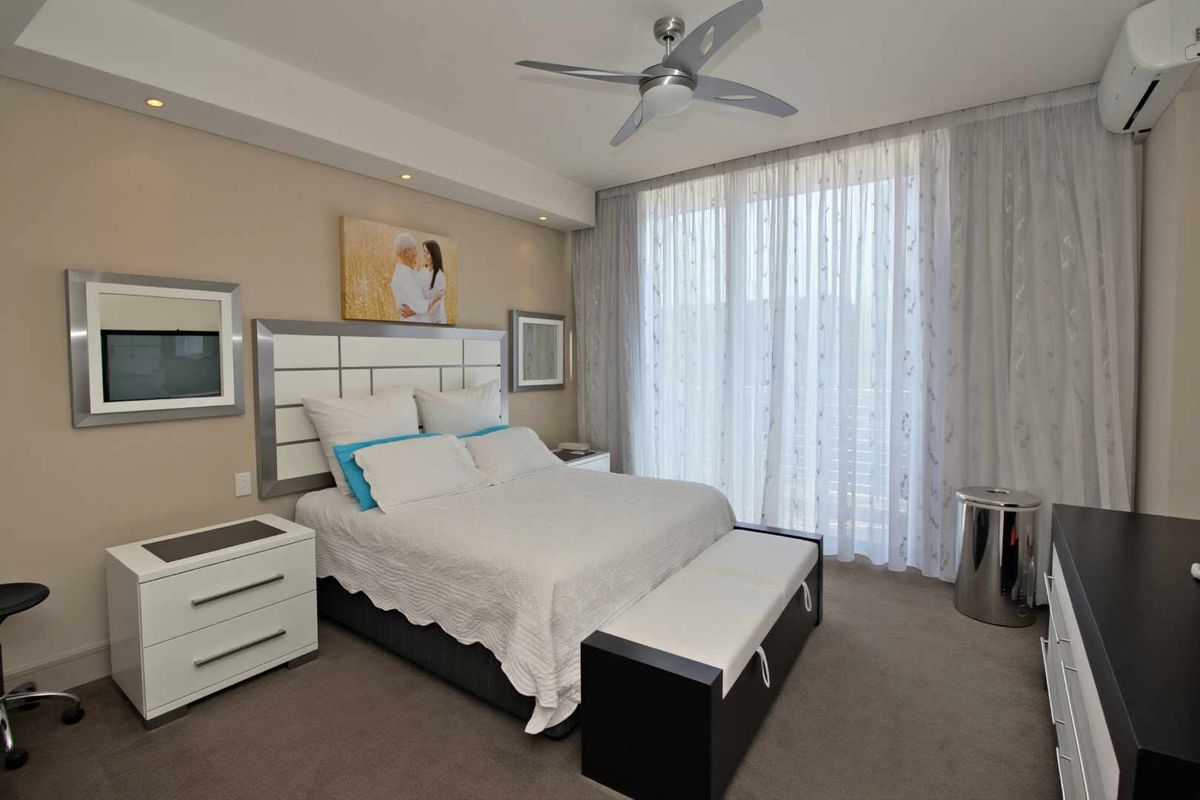 A well-maintained bedroom featuring a ceiling fan, wall-mounted air conditioning unit, and large floor-to-ceiling windows with sheer curtains.