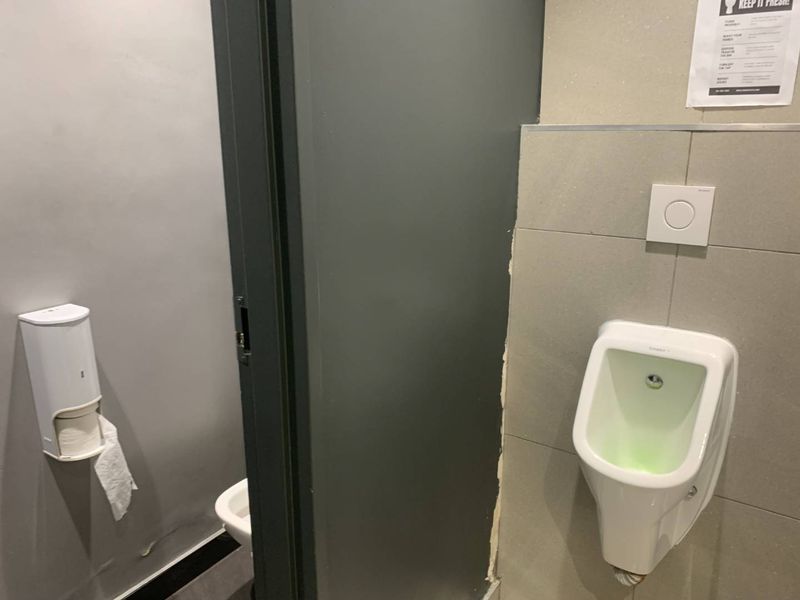 A functional commercial restroom facility featuring a wall-mounted urinal, tiled wall finishes, and a partition door, showing signs of wear and maintenance needs.