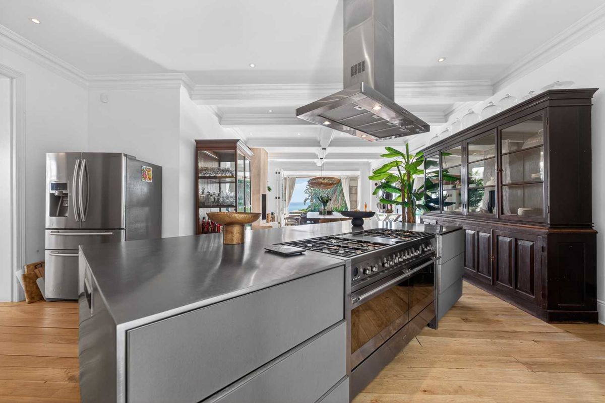 Modern kitchen featuring a large stainless steel island, professional-grade gas range, extractor hood, and elegant hardwood flooring, flowing into an open-plan living area.
