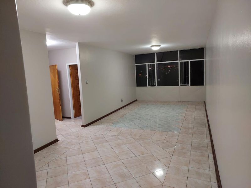 A spacious, empty living area featuring tiled flooring, flush-mount ceiling lighting, and large windows with security bars.