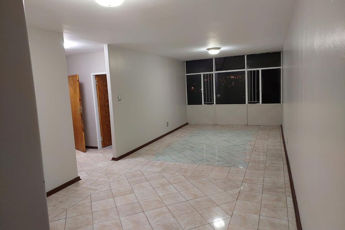 A spacious, empty living area featuring tiled flooring, flush-mount ceiling lighting, and large windows with security bars.
