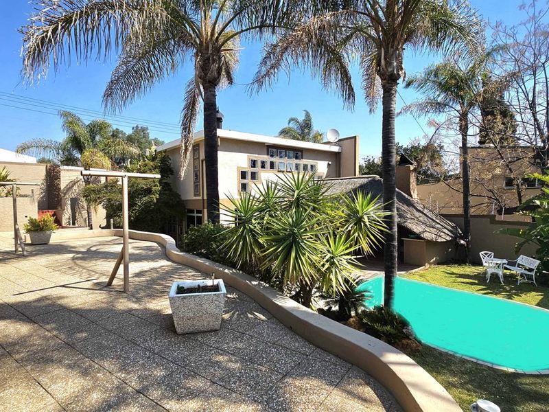 Rear exterior view featuring a paved patio area, swimming pool, and a thatched lapa, set within a landscaped garden with mature palm trees.