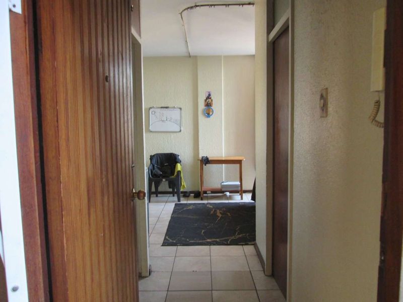A narrow apartment hallway featuring tiled flooring, a solid wood entrance door, and an intercom system, showing signs of wear and exposed ceiling wiring.